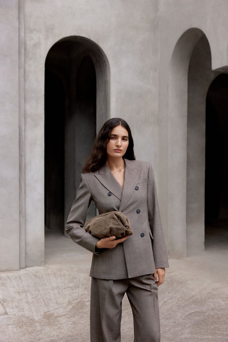 Portrait of a young adult woman in a grey suit holding a taupe clutch bag, standing in front of arches, fashion editorial.