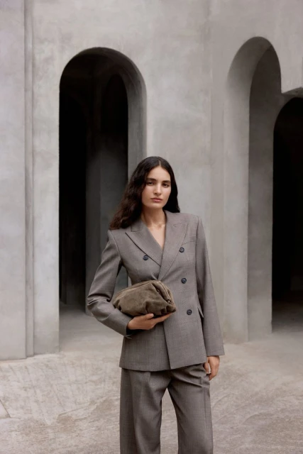 Portrait of a young adult woman in a grey suit holding a taupe clutch bag, standing in front of arches, fashion editorial.