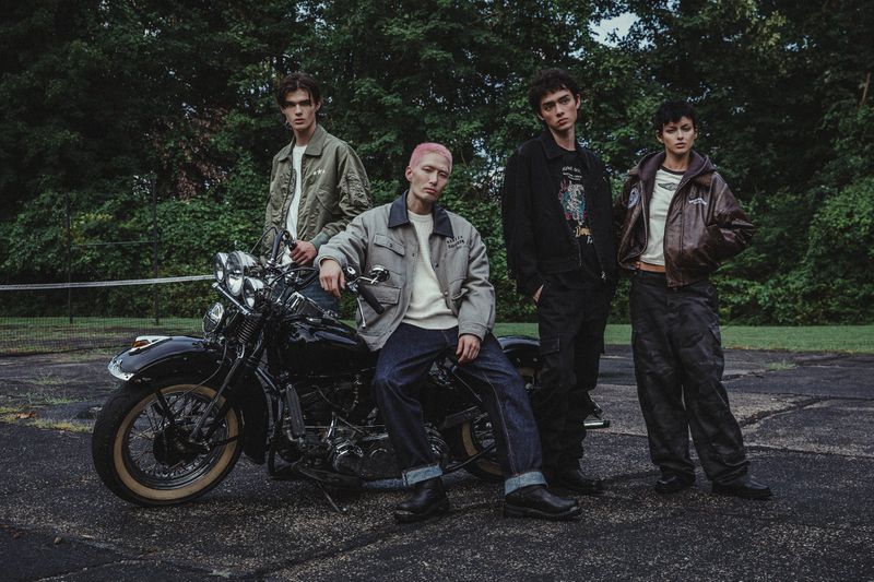 Four young adults pose outdoors next to a classic black motorcycle on asphalt with green trees in the background in an eye-level shot.