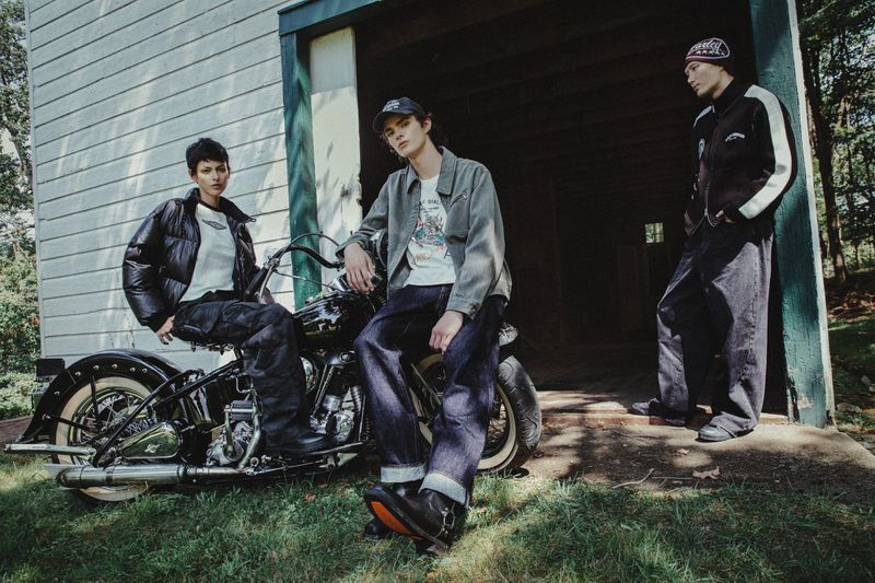 Three fashion models pose with a motorcycle outside a vintage building on a grassy lawn in a medium shot on a sunny day.