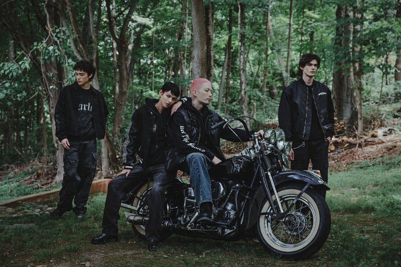 Four fashion models pose with a black motorcycle in a forest setting, wearing dark clothing. The scene has a moody, editorial feel.