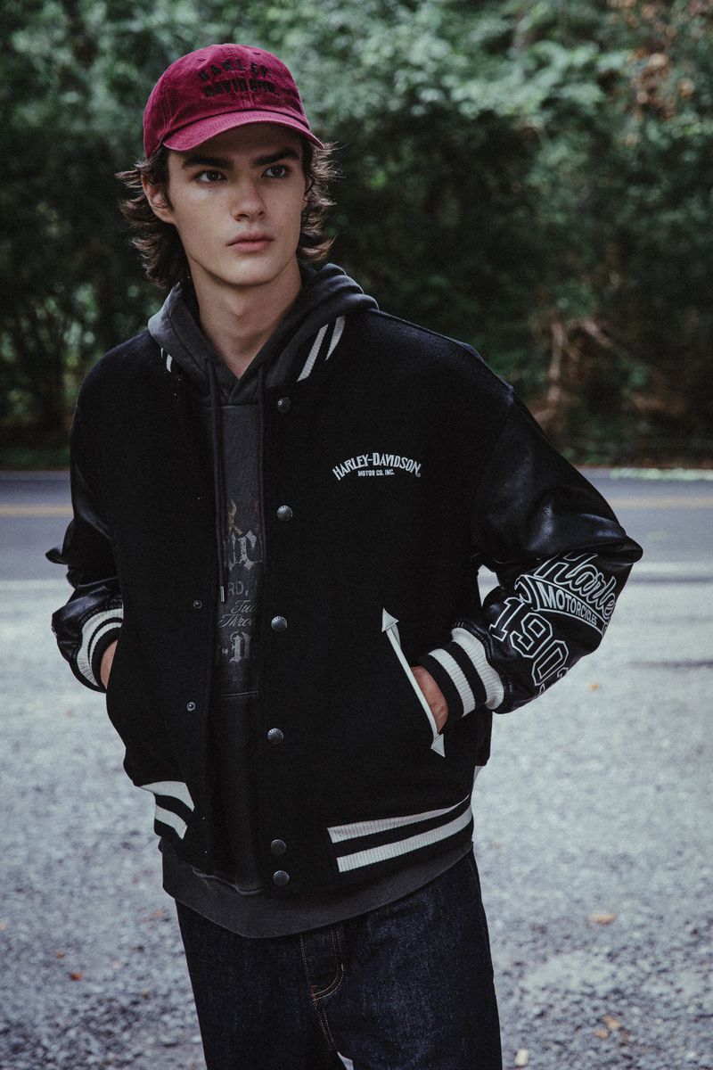 Young adult male in a black Harley-Davidson varsity jacket and cap stands outside with greenery in the background.