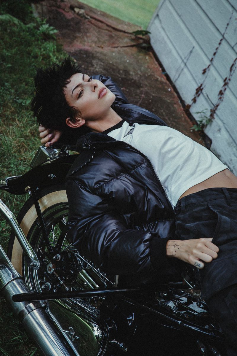 Young woman lies on a vintage motorcycle in a yard with a weathered building; she wears a puffer jacket and dark pants.
