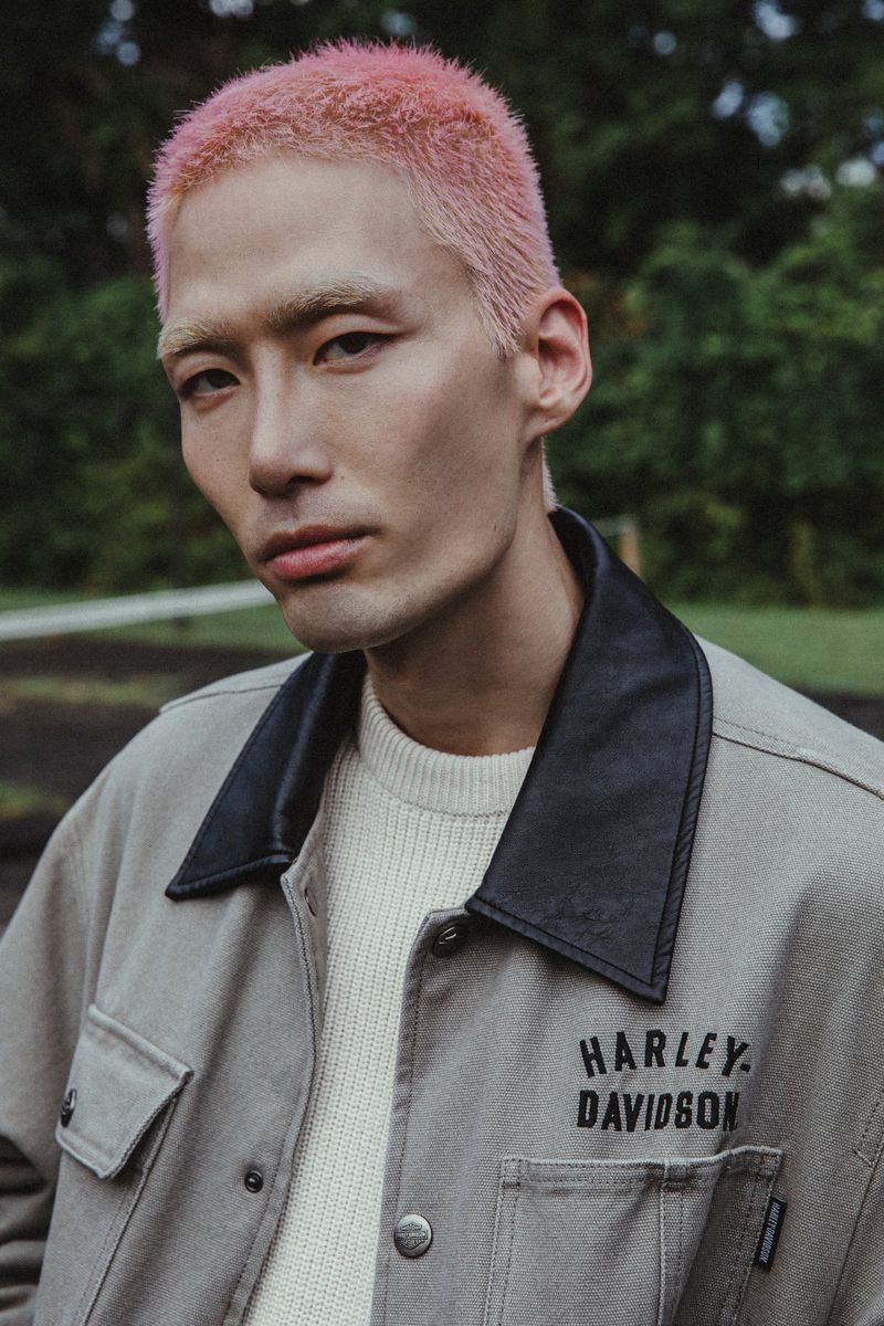 An Asian adult male with pink hair, wearing a cream sweater and Harley Davidson jacket poses in a natural outdoor setting.