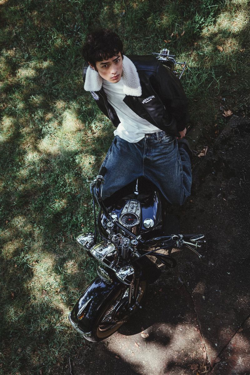 Eye-level shot of a young adult on a motorcycle with a leather jacket, jeans, and white t-shirt, parked on a green grassy surface.