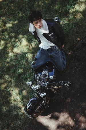 Eye-level shot of a young adult on a motorcycle with a leather jacket, jeans, and white t-shirt, parked on a green grassy surface.