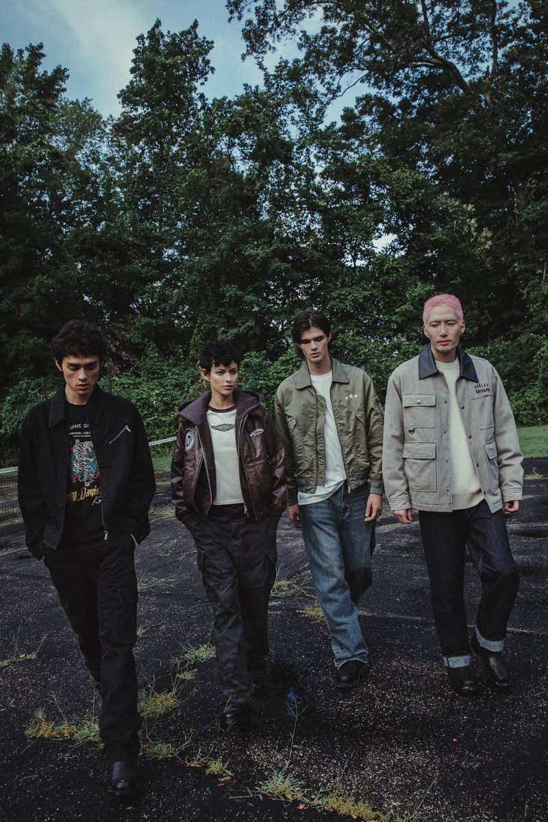 Four young adults in jackets and casual pants walk on cracked asphalt with overgrown trees in the background, captured in a medium shot.