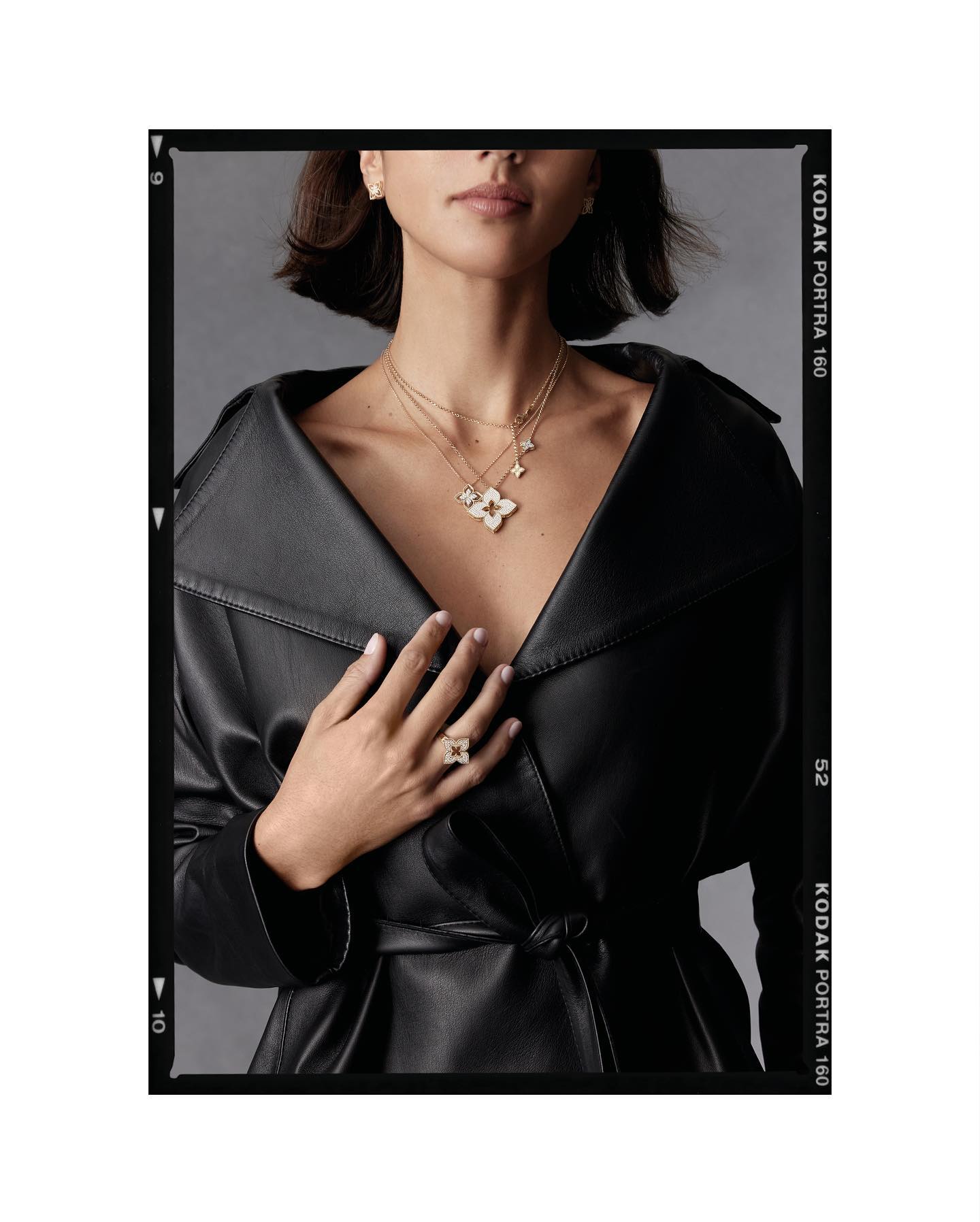 Adult female model in black leather coat with layered necklaces and a ring on her finger poses in a fashion studio setting.