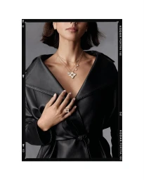 Close-up portrait of a woman wearing layered gold pendants, a statement ring, and a black leather coat against a grey backdrop.