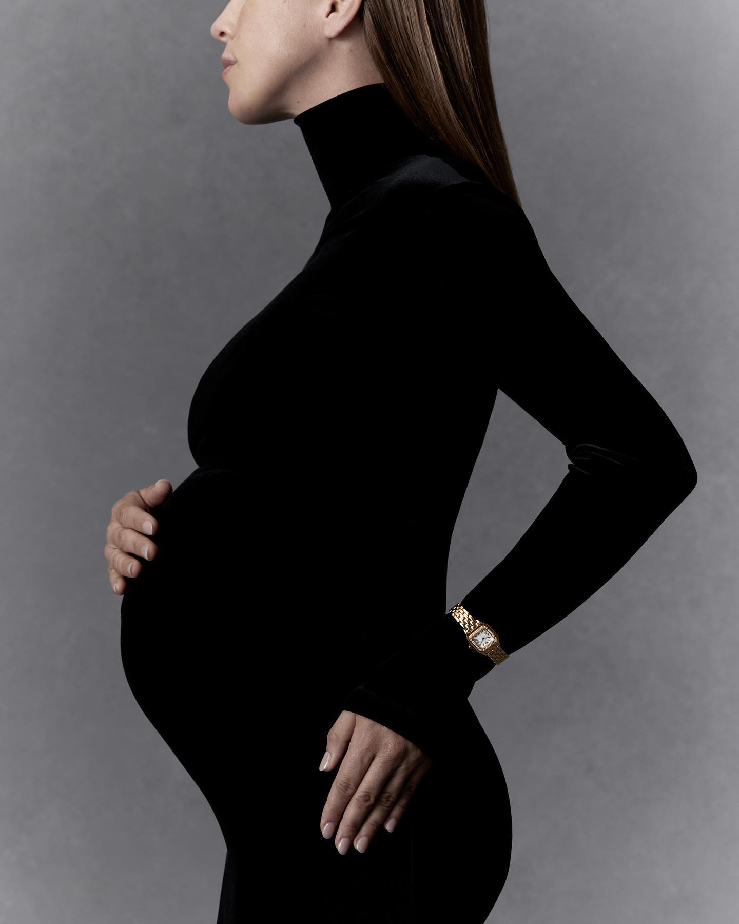Pregnant woman in a black dress, shown in profile, with her hands on her belly and a gold watch, against a soft grey background.