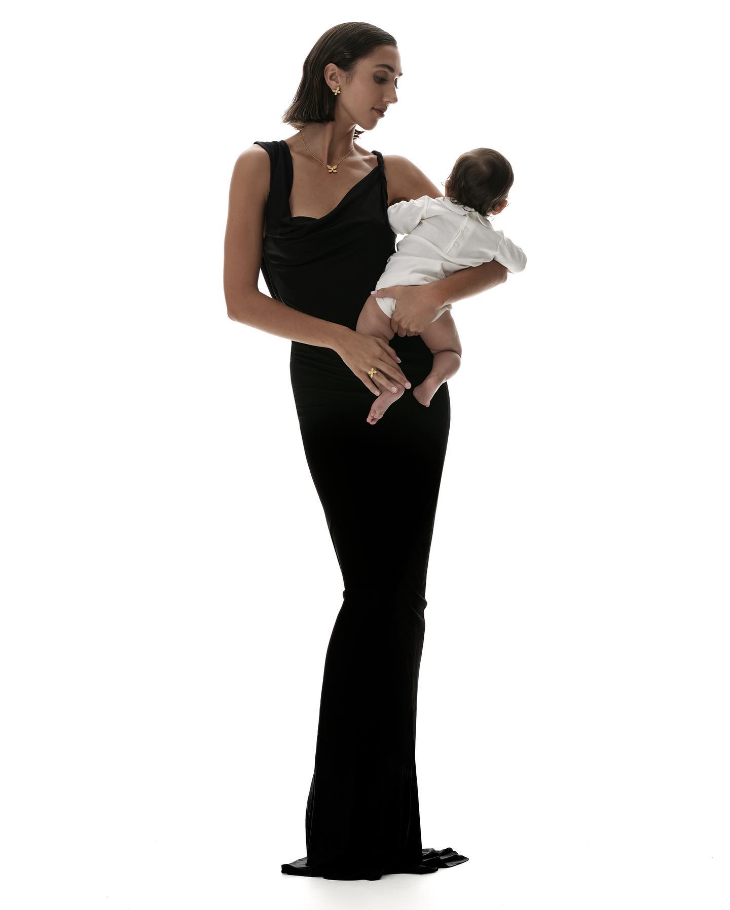 Woman in a long black dress holding an infant in a white shirt against a plain white seamless studio background.