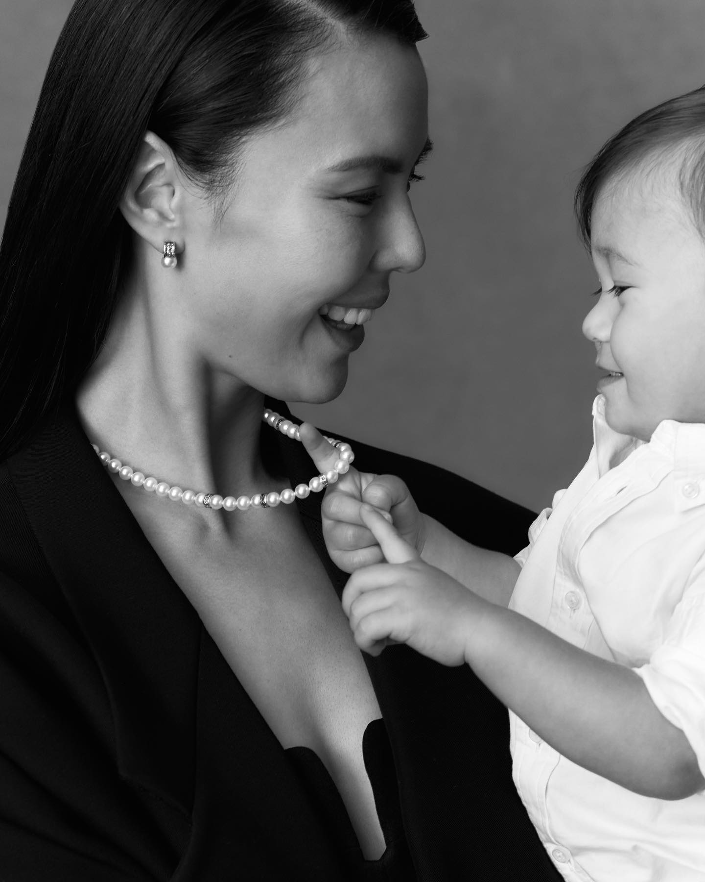Monochrome portrait of a smiling woman wearing a pearl necklace and earrings while holding a child touching the pearls.