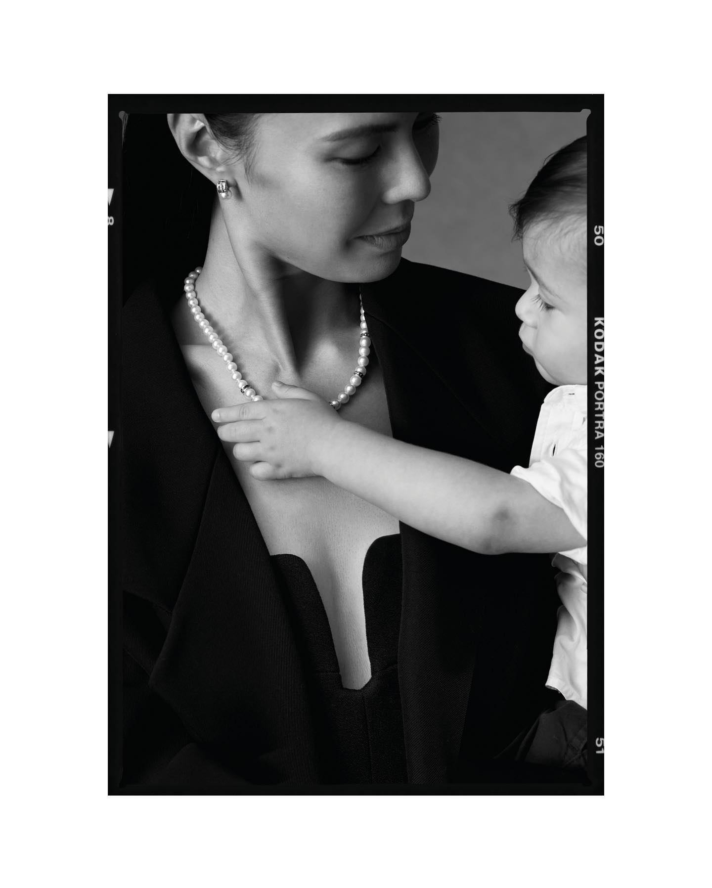 Black and white portrait of a woman wearing a pearl necklace with a child touching the pearls for the She's Extraordinary project by Kennedy.