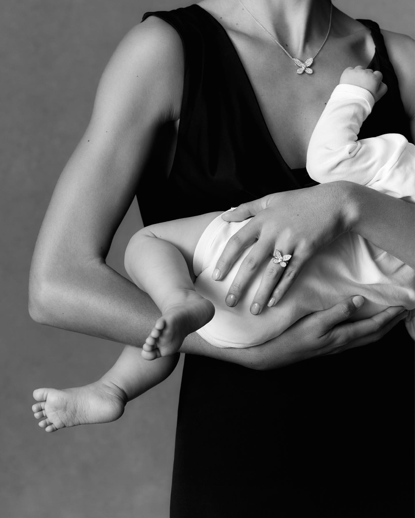 Monochrome image of a woman holding a baby, adorned with butterfly jewelry, against a neutral background.