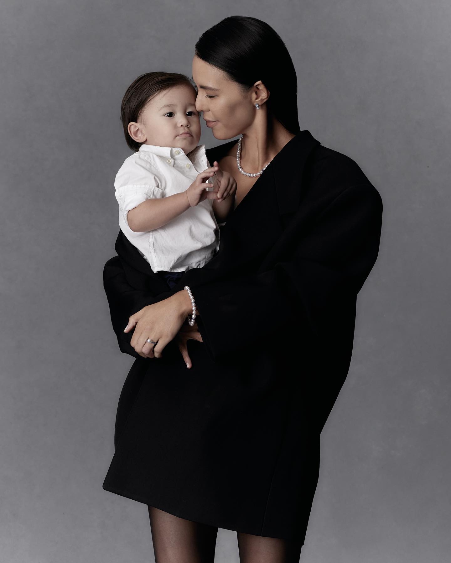 A young child is held by an adult female wearing a black jacket and pearls, set against a gray studio backdrop in a medium shot.