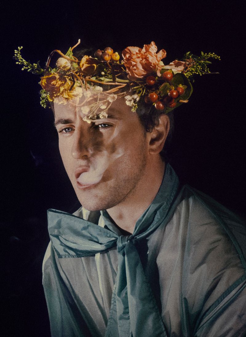 Portrait of a man with a flower crown exhaling vapor, wearing a semi-transparent shirt and neck scarf, set against a dark background.