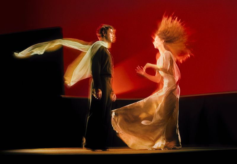 Ballet dancers in motion against a red background. A man in dark clothing and a woman in light clothing are performing a dance.