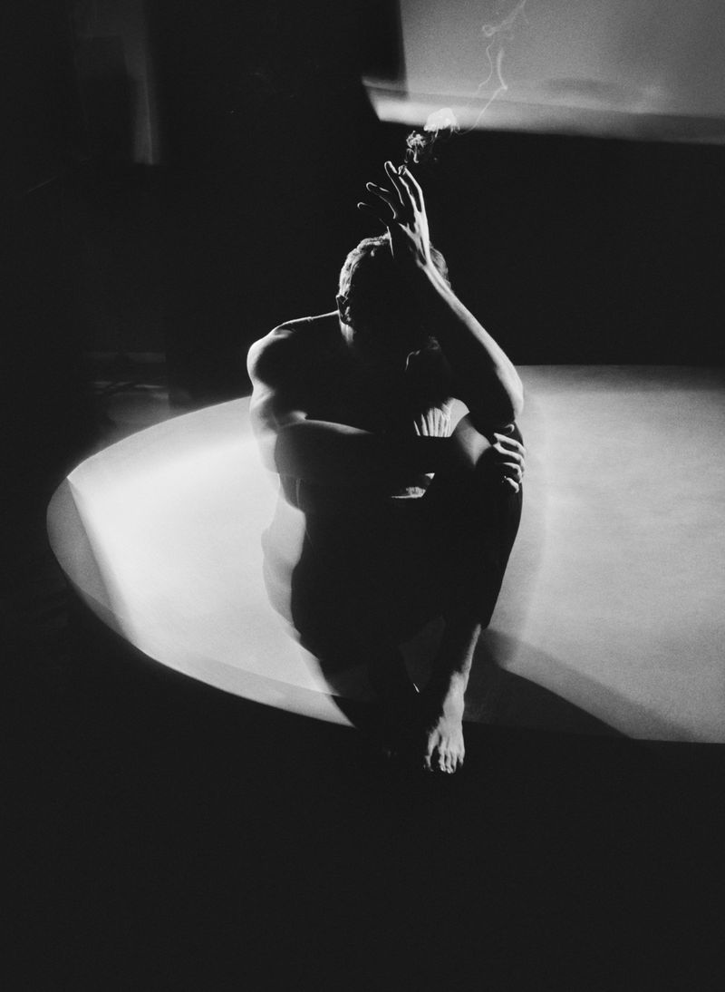 Monochrome photo of shirtless adult male sitting in a dimly lit space with hands raised near his face and a cigarette in his hand.