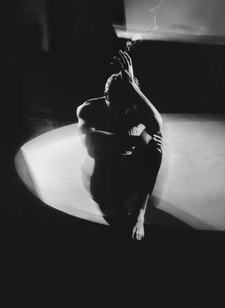 Monochrome photo of shirtless adult male sitting in a dimly lit space with hands raised near his face and a cigarette in his hand.