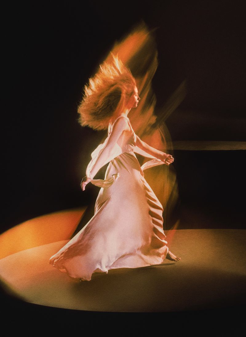 Woman in a flowing white dress dances in a pool of light against a dark background, creating a sense of movement and ethereal beauty.