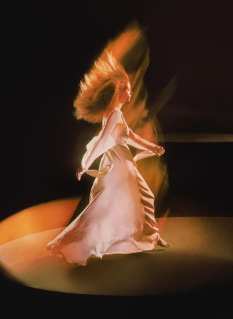 Woman in a flowing white dress dances in a pool of light against a dark background, creating a sense of movement and ethereal beauty.