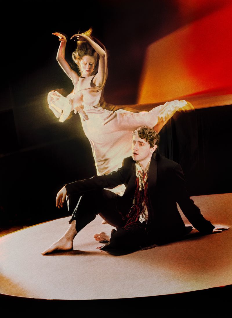 Artistic image features a seated man in a suit alongside a woman in a light-colored dress with motion blur, set against a stage-like backdrop.