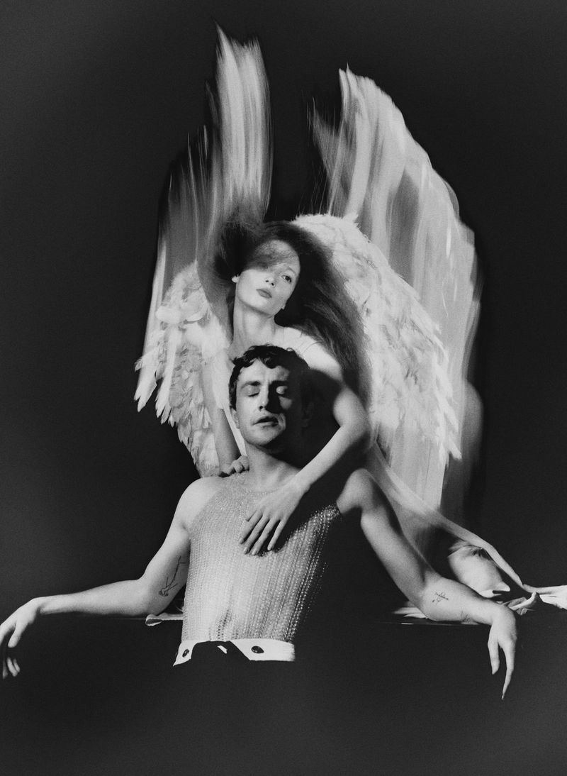 Striking black and white photograph of a man seated with eyes closed, being embraced by a woman with large feathered angel wings.