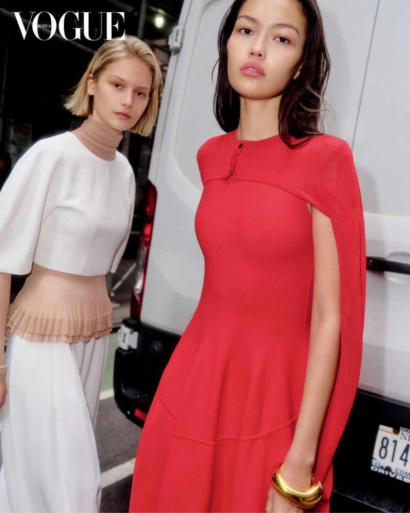 Two fashion models pose outdoors in a medium shot; one wears a red dress and a gold bracelet, the other a white two-piece outfit. Vogue logo.