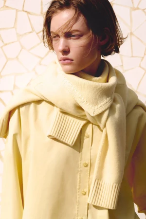 Model Maya Vozhdaeva wearing a pale yellow button-down shirt and draped knit sweater against a mosaic tile wall.
