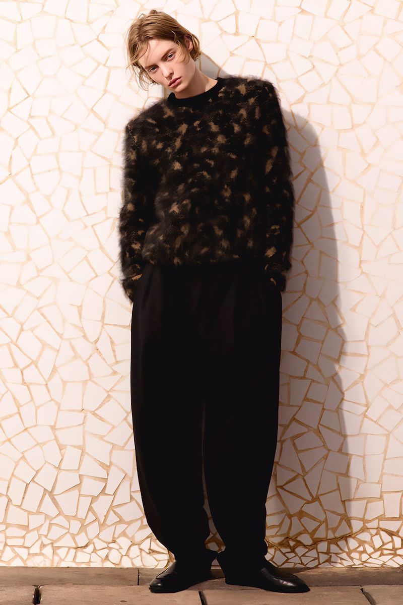 Portrait of a model wearing a furry patterned sweater and baggy black pants, posing against a cracked-tile wall in a studio setting.
