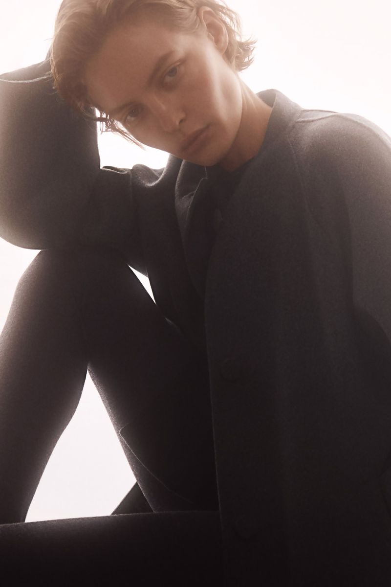 A young adult woman with short hair poses in soft, diffused lighting while wearing a dark, textured, buttoned jacket.