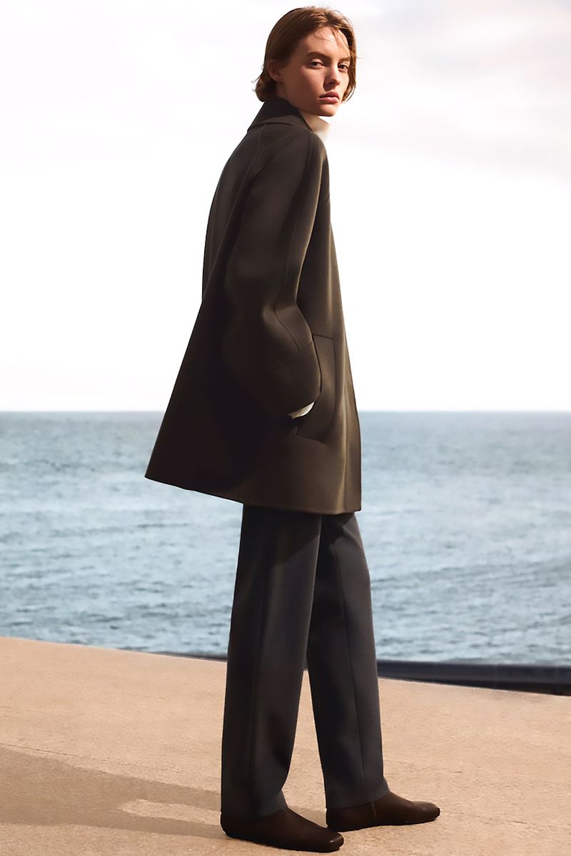 Model Maya Vozhdaeva standing in profile wearing a dark tailored coat and trousers against an open ocean backdrop.