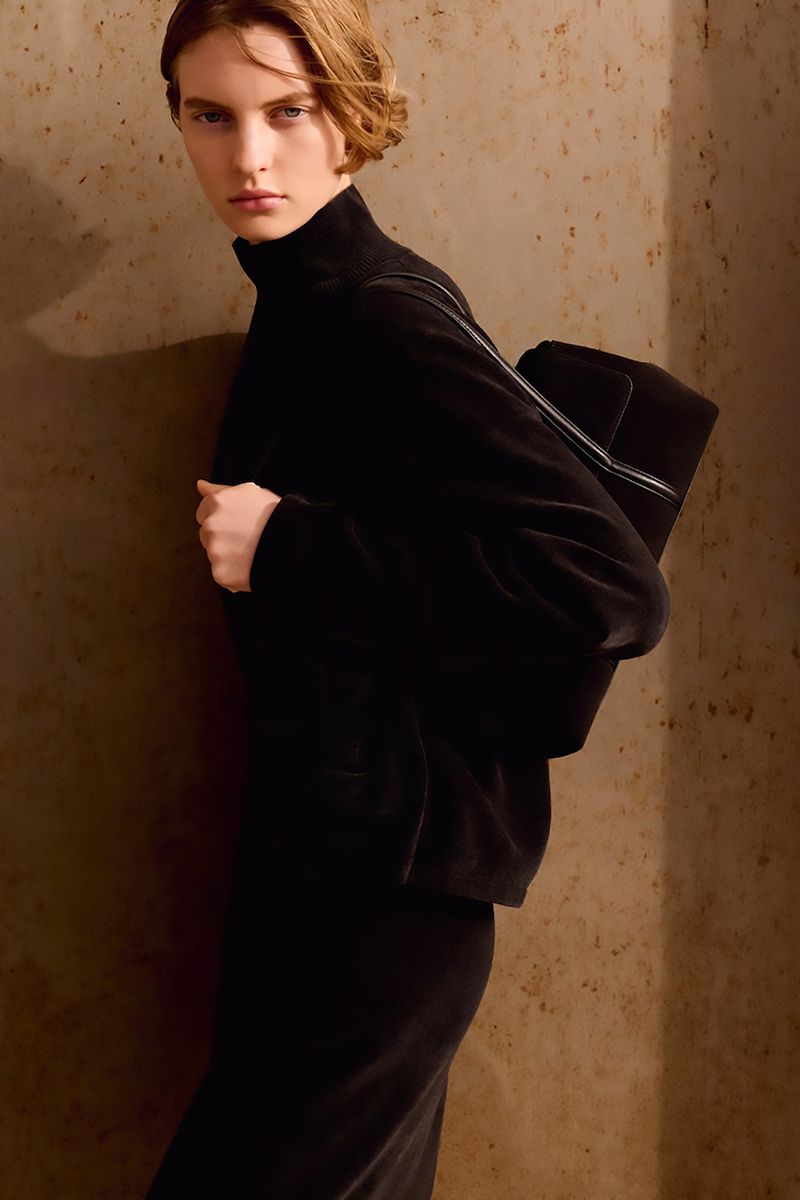 Woman in black turtleneck and skirt with black bag against textured wall, in a fashion editorial style, beauty shot, indoor setting.