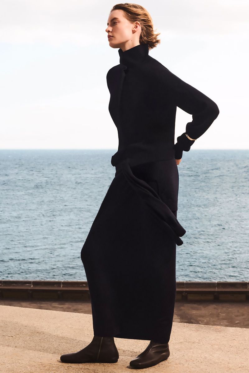A woman stands outdoors wearing a black turtleneck, skirt, and flat shoes, posing near a stone ledge and ocean in the background.