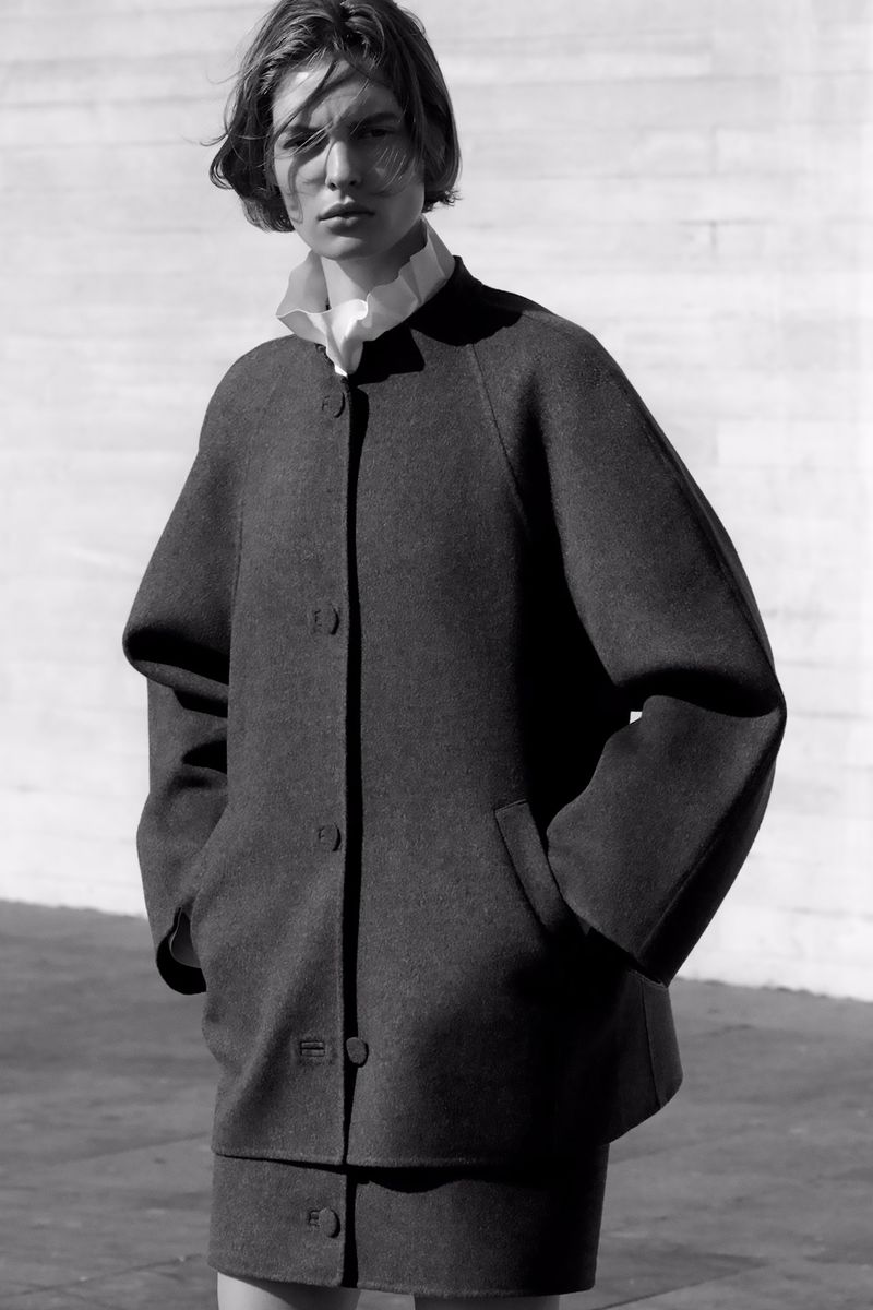 Monochrome fashion portrait of a young woman wearing a dark coat and skirt with a white ruffled collar against a textured wall.