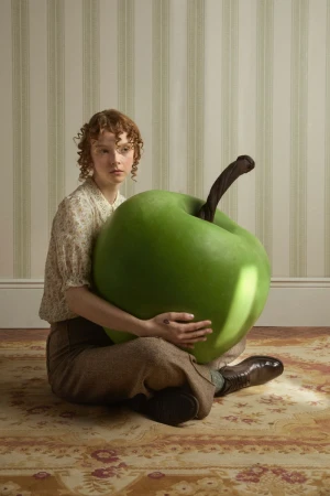 A young woman with curly red hair sits on a patterned rug and hugs a giant, green apple in a room with striped wallpaper.