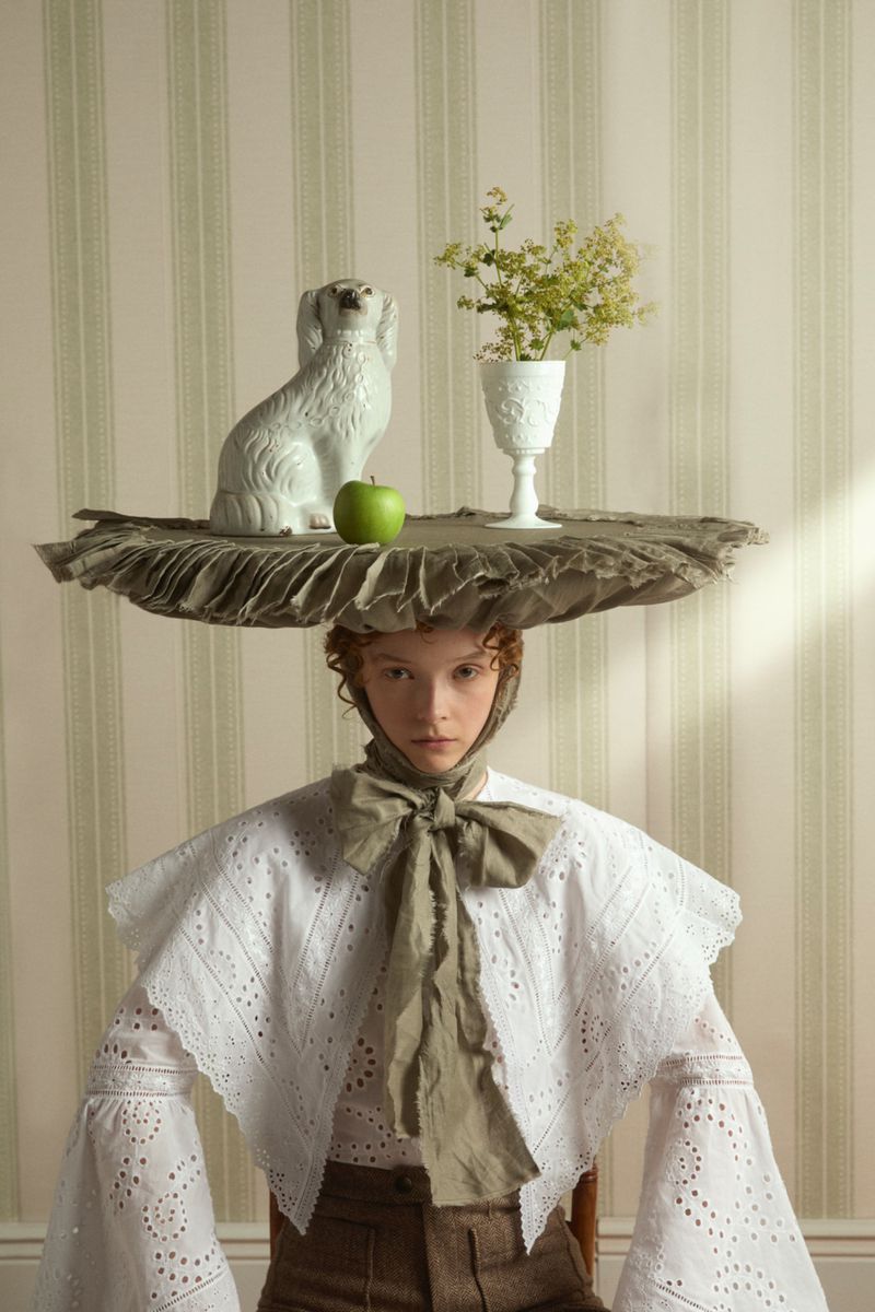 Woman in white blouse with apple, flowers, and dog statue atop her head, posing against a striped wallpaper background in studio.