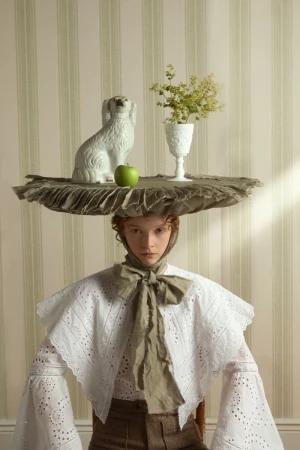 Woman in white blouse with apple, flowers, and dog statue atop her head, posing against a striped wallpaper background in studio.