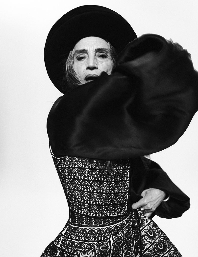 Older woman in black hat and patterned dress poses in a black and white portrait, fashion photography with dramatic lighting.