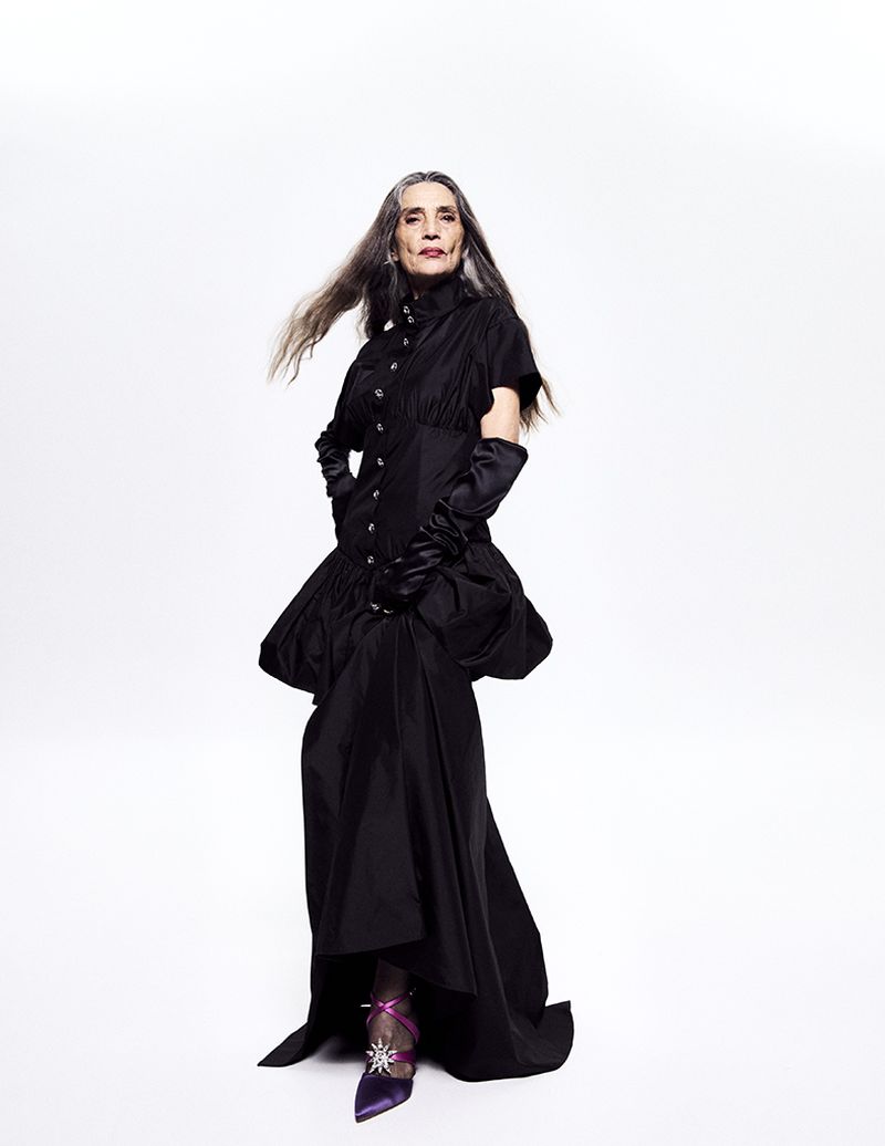 Mature woman poses in a black dress with long gloves against a white background, showcasing a blend of high fashion and elegance.
