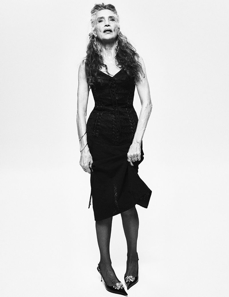 Elegant older woman in a black lace dress and heels posing against a white backdrop in a studio setting. Black and white portrait.