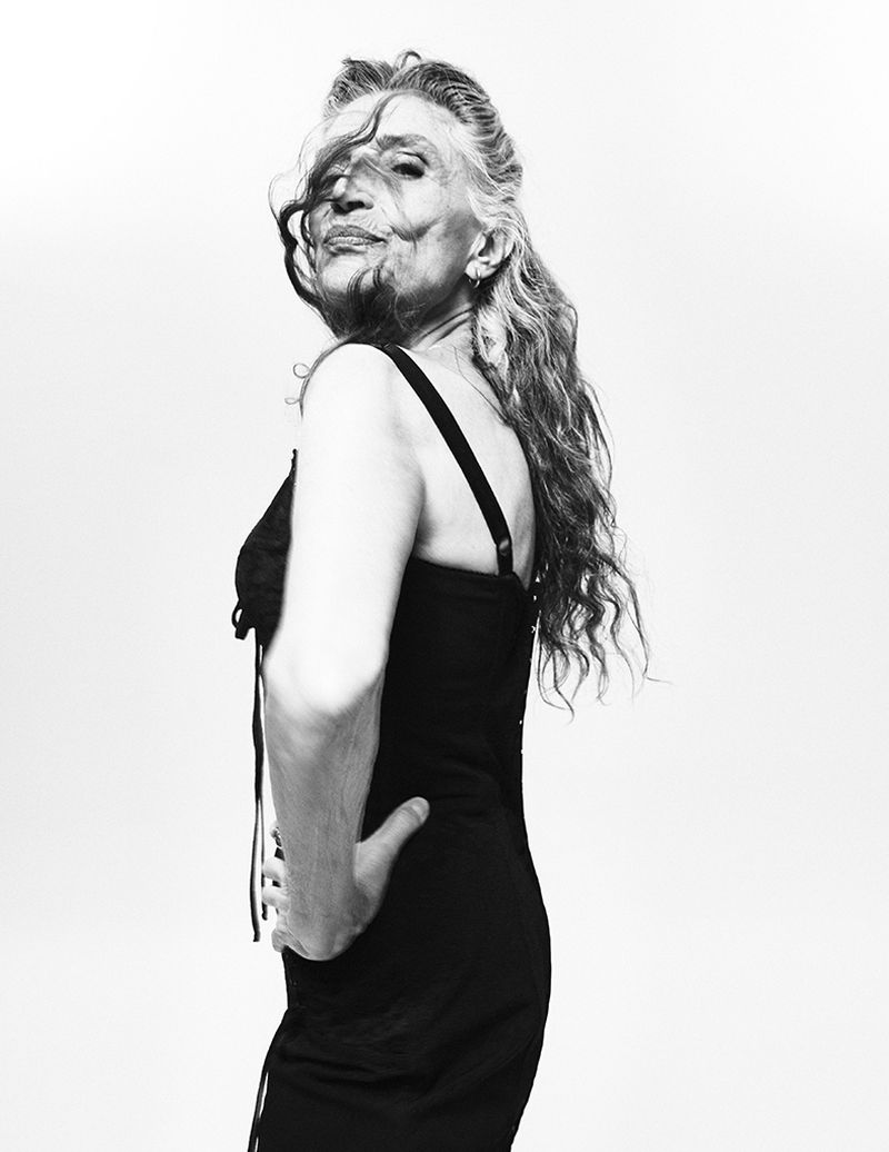Black and white portrait of Ángela Molina in a black slip dress looking over her shoulder against a white studio background.