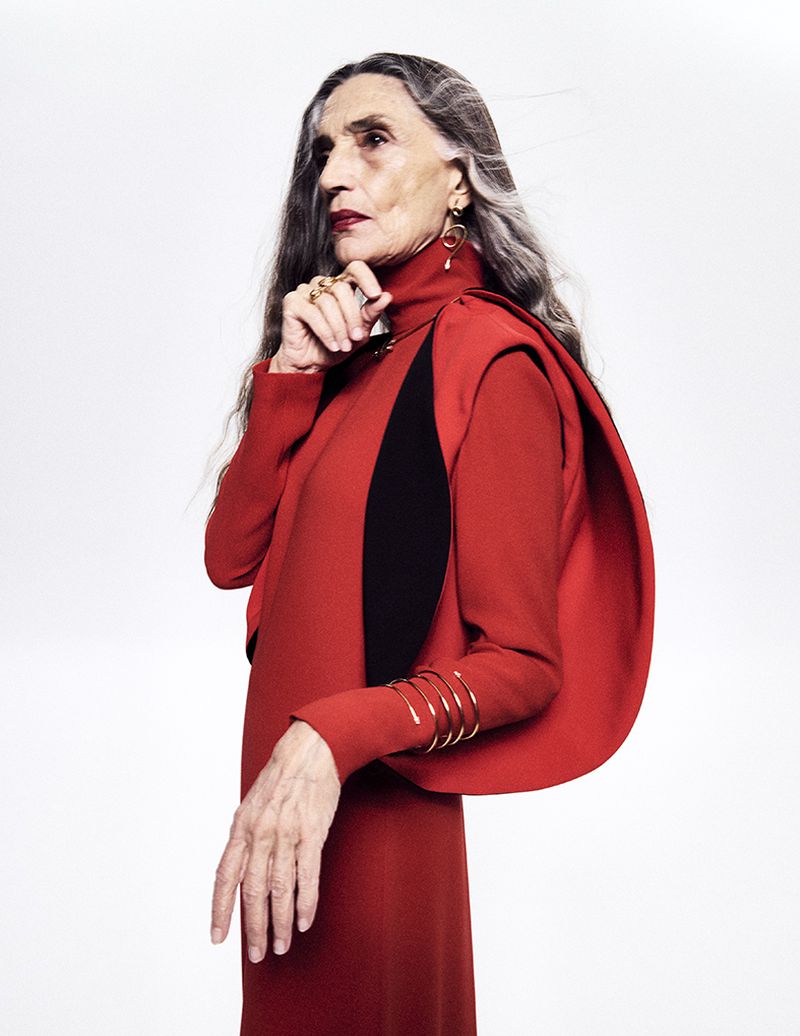 Older woman in a red dress with black accents posing in front of a white background, wearing jewelry and bracelets.