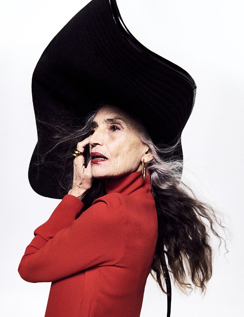 Elegant elderly woman in red turtleneck and large black hat strikes a pose against a bright white background. Fashion editorial.