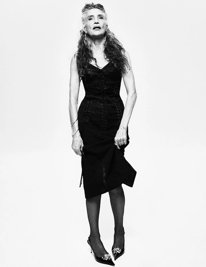 Ángela Molina wearing a black midi dress, sheer tights, and embellished pointed heels against a white studio background.