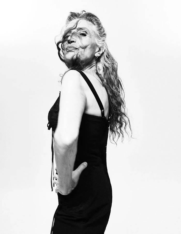 Black and white portrait of Ángela Molina in a black slip dress looking over her shoulder against a white studio background.