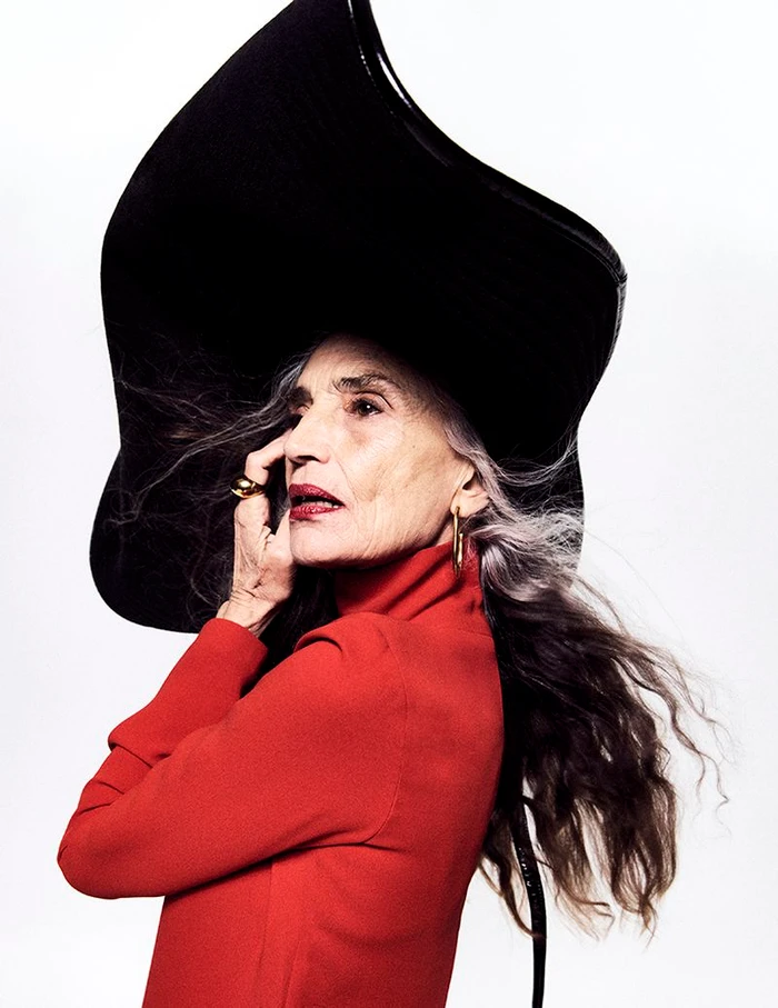 Ángela Molina wearing an oversized black sculptural hat and a red turtleneck top against a bright white background.