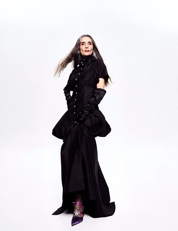 Spanish actress Ángela Molina posing in a black high-collar gown with long satin gloves and purple heels against a white backdrop.