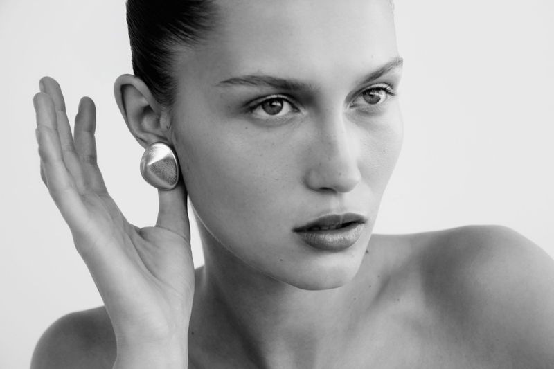 Monochrome close-up of a fair-skinned woman with slicked-back hair, red lipstick, and a large silver earring, set against a white background.