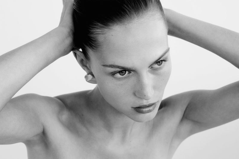 Close-up portrait in black and white of an adult woman with her hands behind her head, looking to the side with a serious expression.